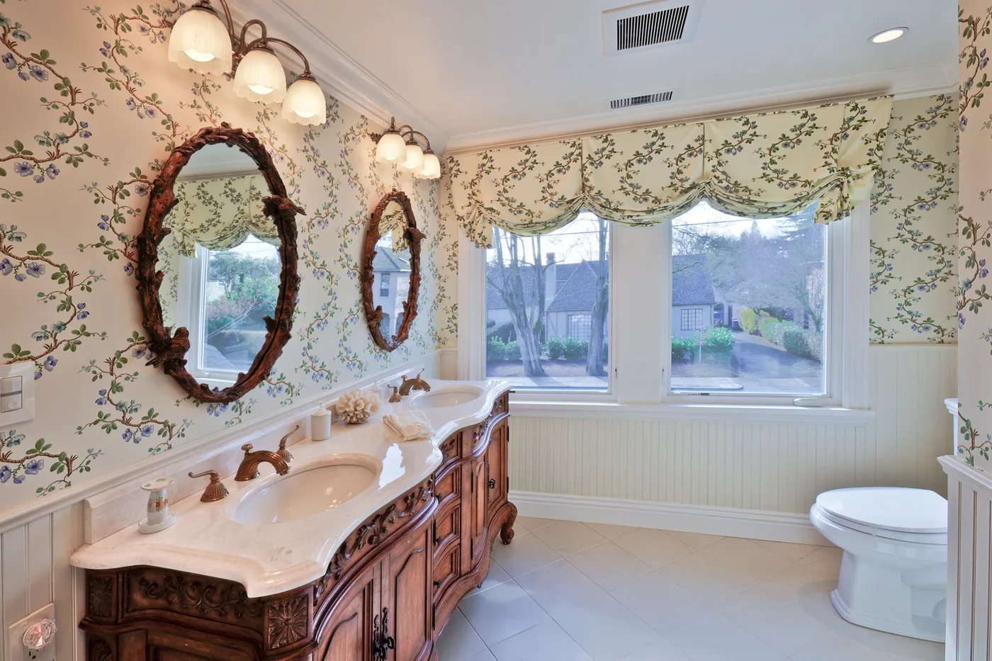 Second bath with a carved double vanity, paired oval mirrors and floral wallpaper