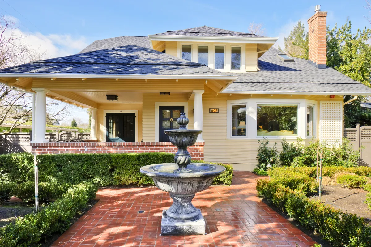 The yellow Craftsman house behind a brick forecourt and a stone fountain, deep eaves over the porch