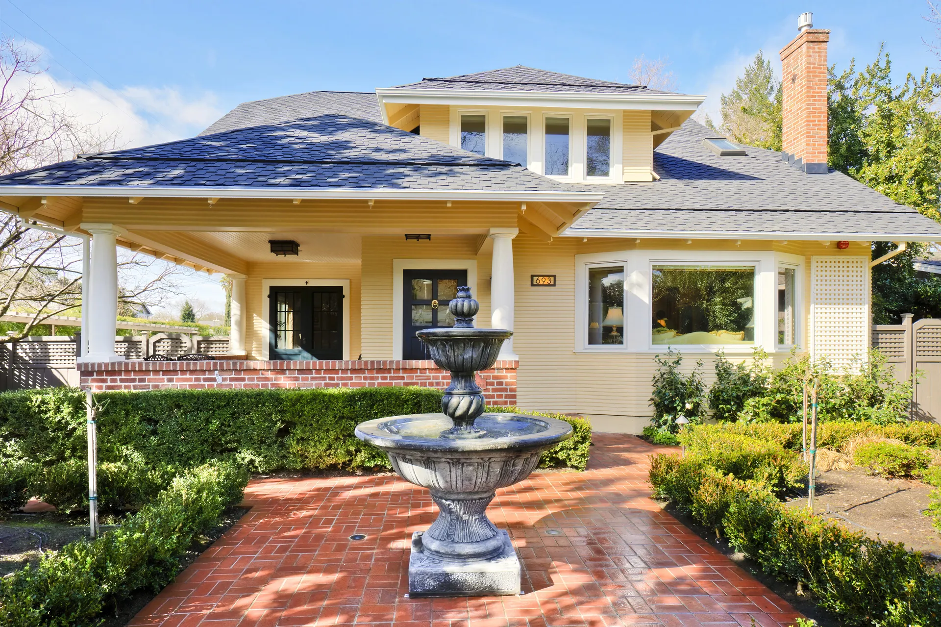 The yellow Craftsman house behind a brick forecourt and a stone fountain, deep eaves over the porch