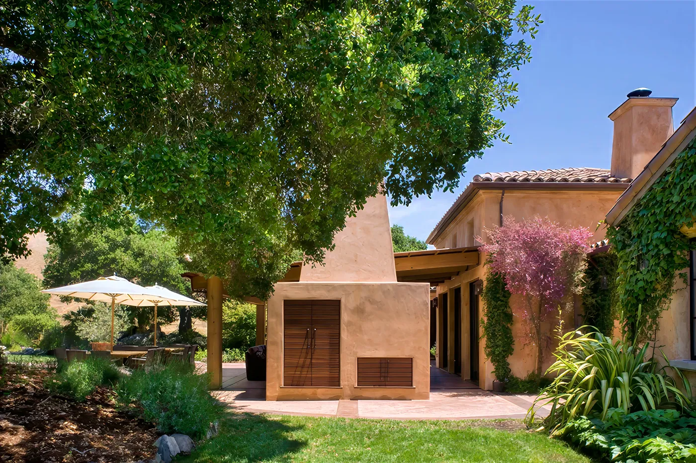 The plaster outdoor fireplace and loggia seen from the lawn, beneath an oak