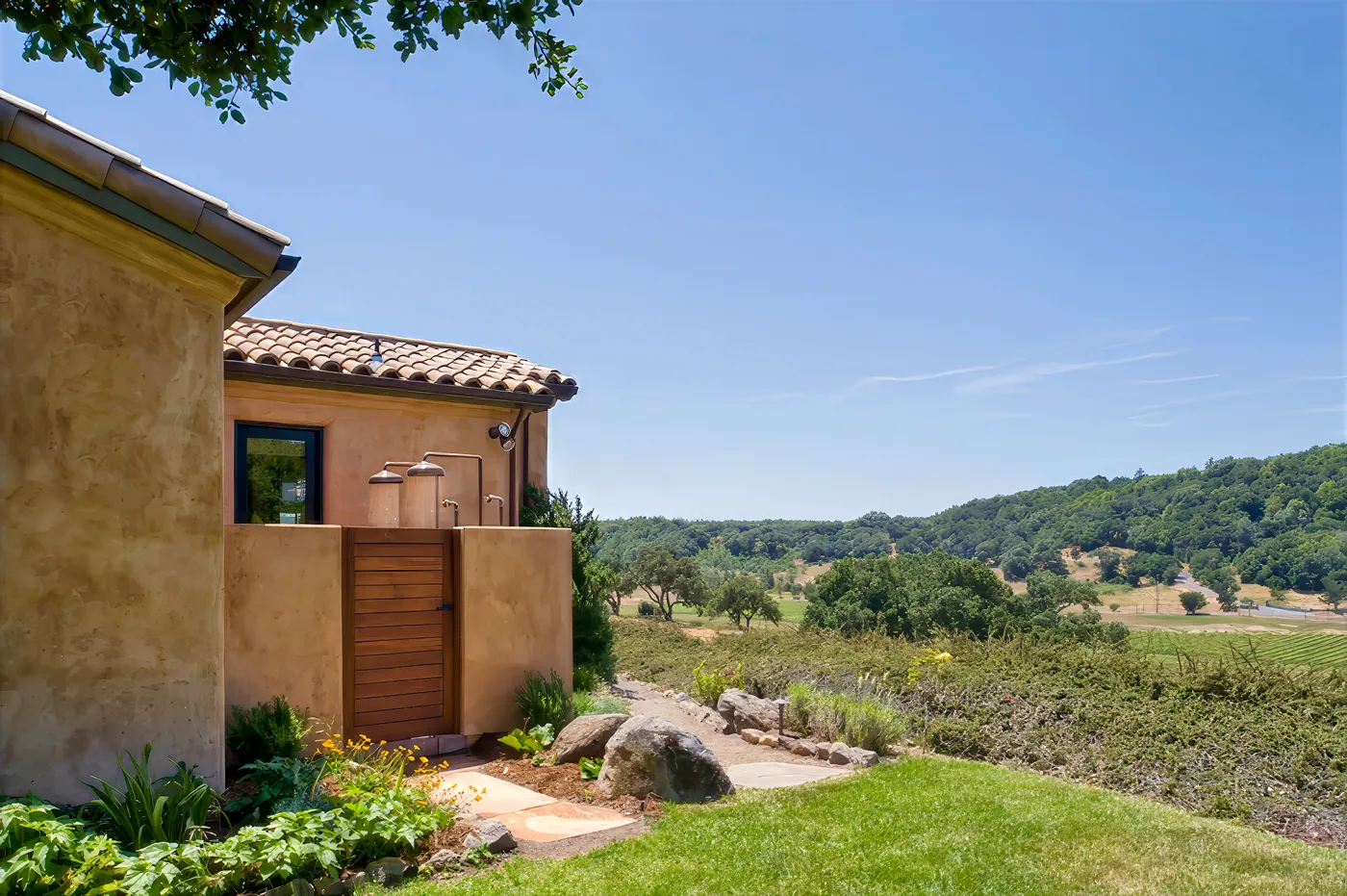 Outdoor shower with twin heads on a plaster wall behind a slatted gate, valley beyond