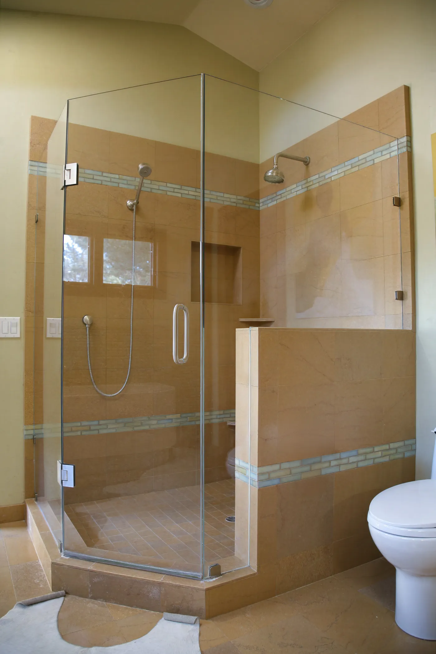 Corner shower in stone tile behind frameless glass, banded with glass mosaic