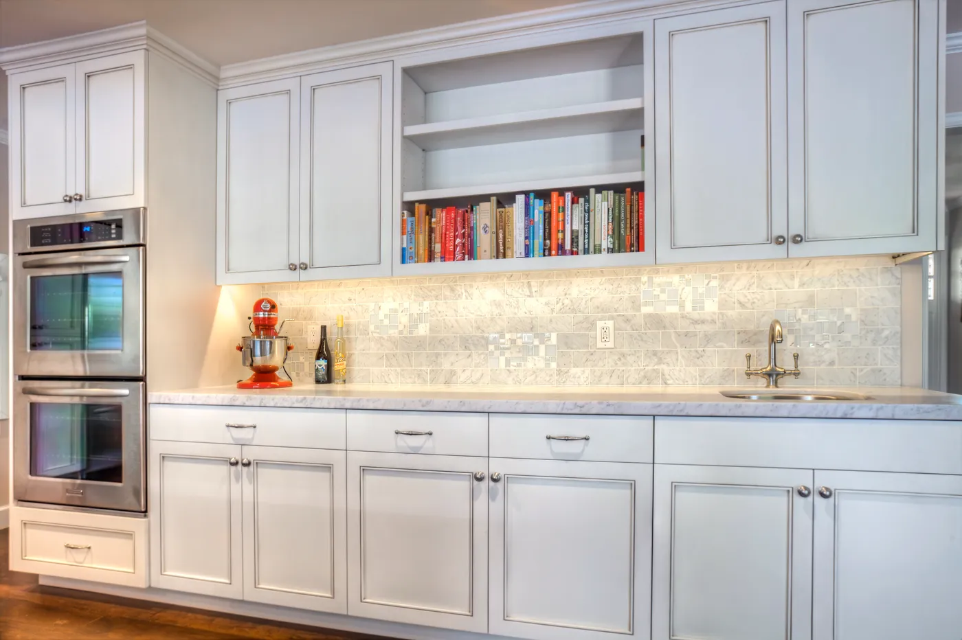 Run of cabinetry with double wall ovens, an open cookbook shelf and a marble counter