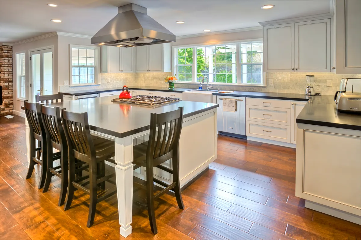 Kitchen island with a stainless hood above it and stools along one side, white cabinetry beyond