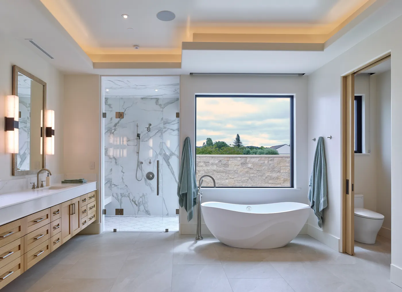 Freestanding tub below the window in the primary bath, beside the marble shower
